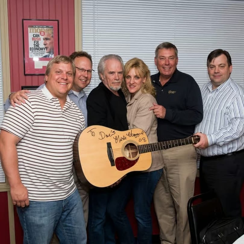 Six people, including Merle Haggard, pose with an autographed guitar in a restaurant setting.