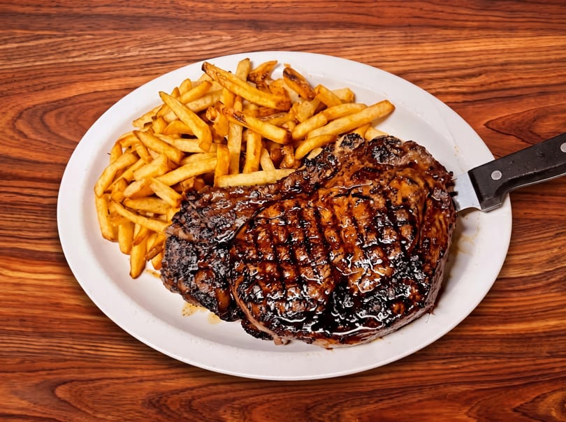 Grilled steak and golden french fries on a white plate with a knife, set on a wooden table.