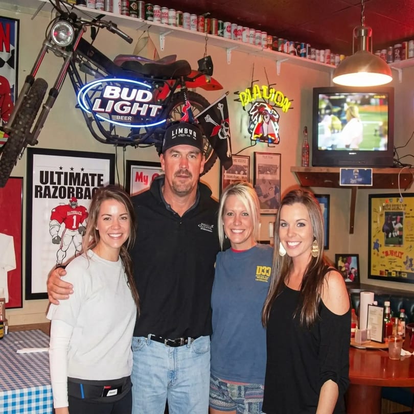 Four smiling people pose in a casual restaurant with sports memorabilia and neon signs.