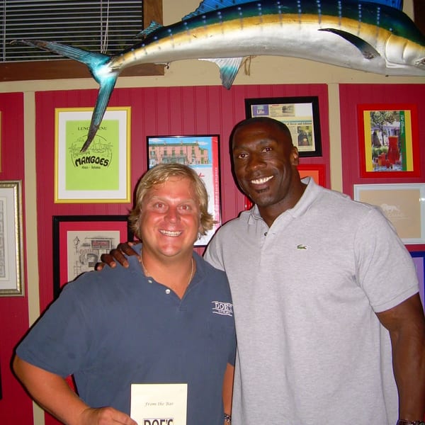 Two smiling men in a restaurant, one holding a "Doe's Eat Place" menu, with a large fish sculpture.