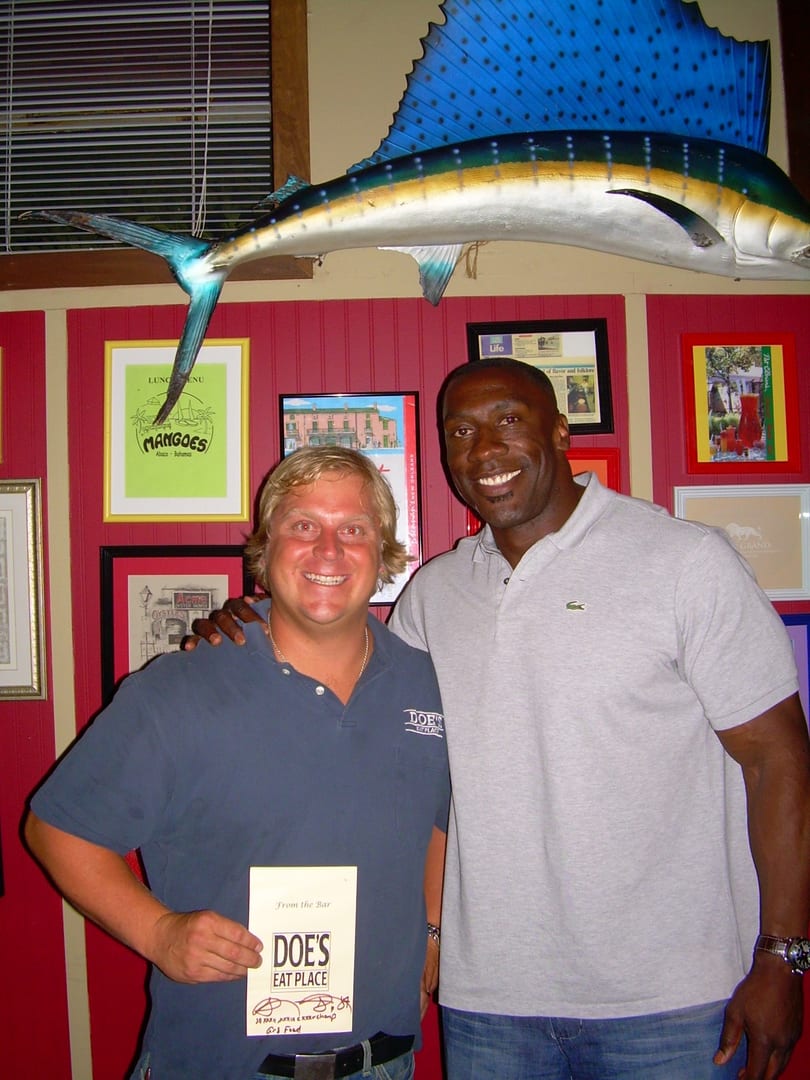 Two smiling men in a restaurant, one holding a "Doe's Eat Place" menu, with a large fish sculpture.