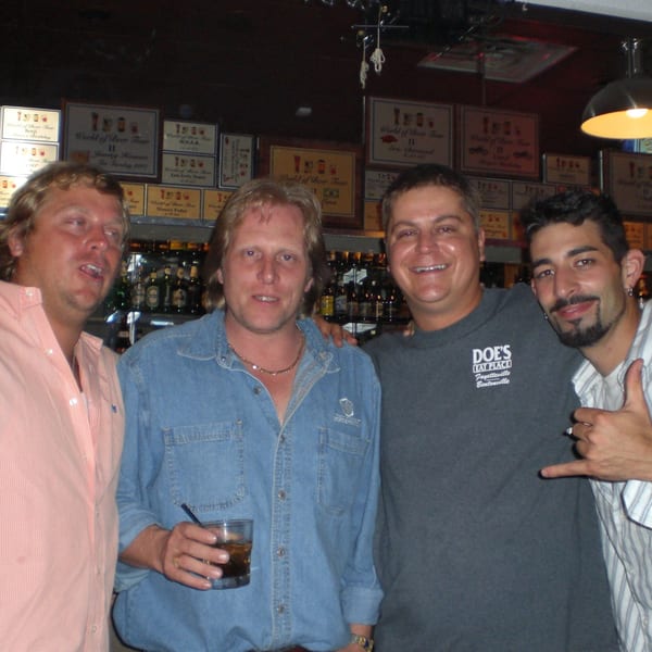 Four smiling men pose in a bar with bottles and plaques, one holding a drink.