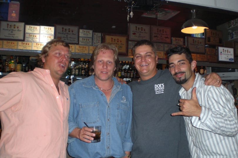Four smiling men pose in a bar with bottles and plaques, one holding a drink.