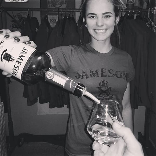 Smiling bartender wearing a Jameson shirt pours whiskey into a glass.